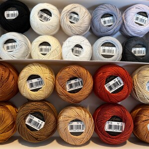 May include: A box of 18 spools of embroidery floss in various shades of brown, beige, and orange. The spools are labeled with the brand name "Anchor" and the number "8".