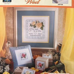 Könnte beinhalten: Ein Kreuzstich-Hochzeitssampler mit dem Text "I Thee Wed" und "Two shall be as One". Das Design umfasst gelbe Rosen und die Namen Angela und Albert mit dem Datum 16. September 2000. Gezeigt werden auch kleine gerahmte Kreuzstichmuster und ein Kissen.