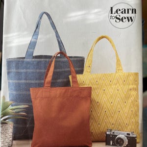 Puede incluir: Patrón de costura Simplicity S9308 para tres bolsos tote. El sobre del patrón muestra un bolso azul a rayas, un bolso liso color óxido y un bolso amarillo con patrón de chevrón blanco. El texto incluye Simplicity, S9308 y Learn to Sew.