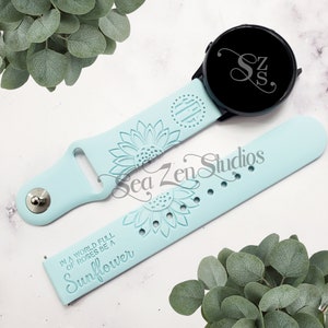 May include: A light blue silicone watch band with a sunflower design and the text "In a world full of roses be a sunflower". The band is compatible with a smartwatch with a black face and a monogrammed initial.