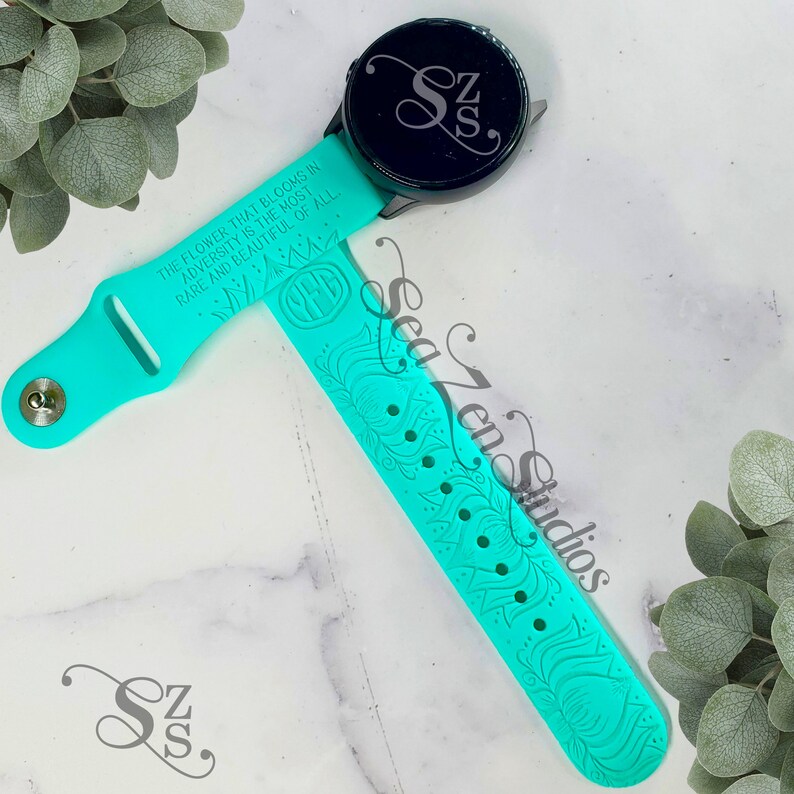 Może przedstawiać: Turkusowy pasek do zegarka wykonany z silikonu z kwiatowym wzorem i tekstem "THE FLOWER THAT BLOOMS IN ADVERSITY IS THE MOST RARE AND BEAUTIFUL OF ALL". Pasek jest kompatybilny z zegarkiem smartwatch z czarną tarczą.