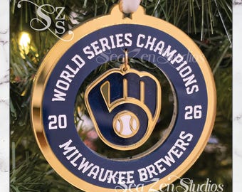 May include: A circular gold and navy blue ornament featuring the text World Series Champions 2026 Milwaukee Brewers. A central navy blue baseball glove icon with a white baseball is suspended within the center of the ring.