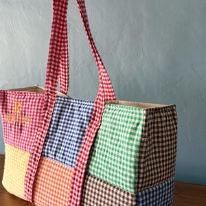 Puede incluir: Un bolso tote rectangular confeccionado con cuadrados de tela acolchada en varios patrones de cuadros vichy en rojo, amarillo, naranja, azul, verde y marr&oacute;n. El bolso tiene asas de cuadros rojos y blancos y el texto bordado R BRIDE.