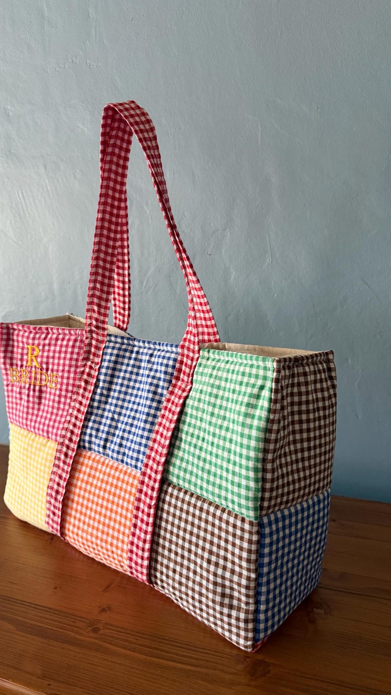 Puede incluir: Un bolso tote rectangular confeccionado con cuadrados de tela acolchada en varios patrones de cuadros vichy en rojo, amarillo, naranja, azul, verde y marr&oacute;n. El bolso tiene asas de cuadros rojos y blancos y el texto bordado R BRIDE.