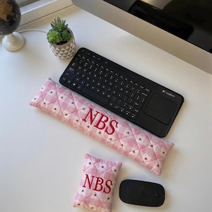 May include: A pink and white quilted keyboard wrist rest and small pillow with the letters "NBS" in red. A black wireless keyboard and mouse are also present. The wrist rest and pillow have a heart pattern.