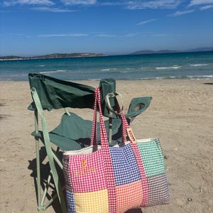 Puede incluir: Un bolso tote rectangular grande con un dise&ntilde;o de patchwork de cuadros vichy multicolores en rojo, azul, verde, amarillo y naranja. El bolso tiene dos asas largas de tela y la letra R bordada con hilo dorado en la secci&oacute;n roja.