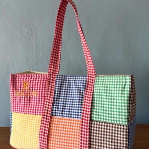Peut inclure: Un sac fourre-tout rectangulaire composé d'un patchwork de carrés de tissu vichy rouge, bleu, vert, jaune, orange et marron. Le sac présente des anses en vichy rouge et blanc et le texte brodé en jaune R BRIDE sur la section rouge.
