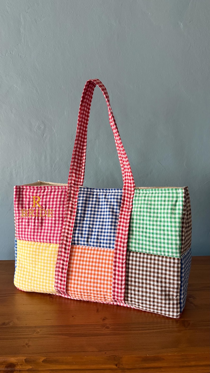 Puede incluir: Un bolso tote rectangular con un dise&ntilde;o de patchwork de cuadros vichy multicolores en rojo, azul, verde, amarillo, naranja y marr&oacute;n. El bolso tiene dos asas de cuadros vichy rojos y blancos y el texto bordado en amarillo R BRIDE al frente.
