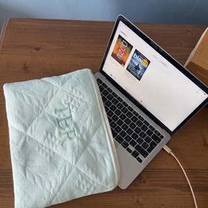 May include: A light mint green quilted laptop sleeve featuring the embroidered letters TBR on the front, positioned next to an open silver laptop displaying a digital library interface with book covers on a wooden surface.
