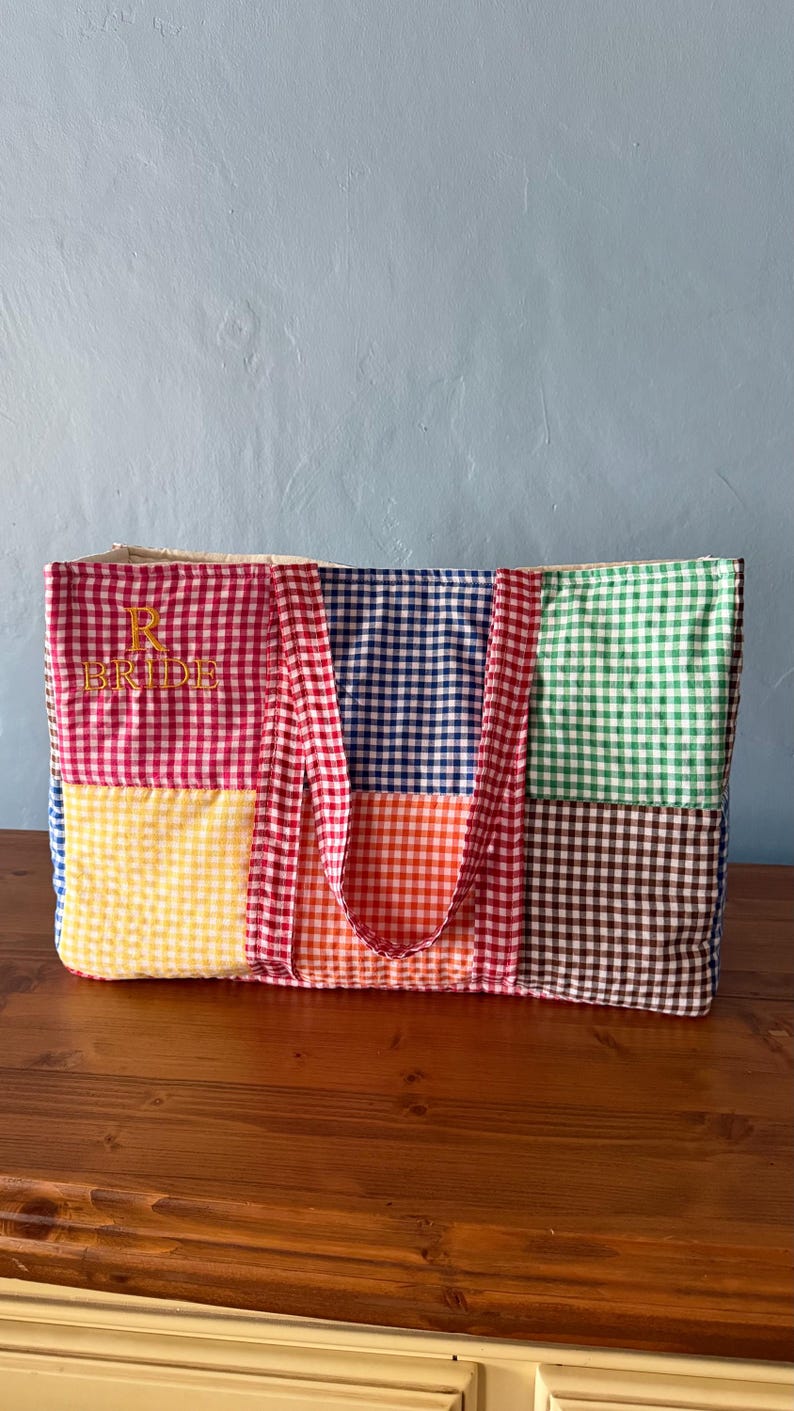 Puede incluir: Un bolso tote rectangular confeccionado con cuadrados de tela acolchada con un patr&oacute;n de cuadros vichy multicolor en rojo, amarillo, azul, naranja, verde y marr&oacute;n. La secci&oacute;n de cuadros rojos incluye el texto amarillo bordado R BRIDE.