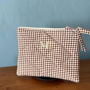 Può includere: Una pochette trapuntata rettangolare con un motivo a quadretti vichy marroni e bianchi. La pochette presenta una chiusura a zip color crema con una linguetta in tessuto abbinata. Al centro è ricamato il monogramma MF in filo dorato.