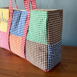Puede incluir: Un bolso tote rectangular hecho de paneles de tela acolchada con un patr&oacute;n de cuadros vichy multicolor en amarillo, azul, naranja, verde y marr&oacute;n. El bolso tiene dos asas de cuadros vichy rojos y blancos y el texto I ROOM.