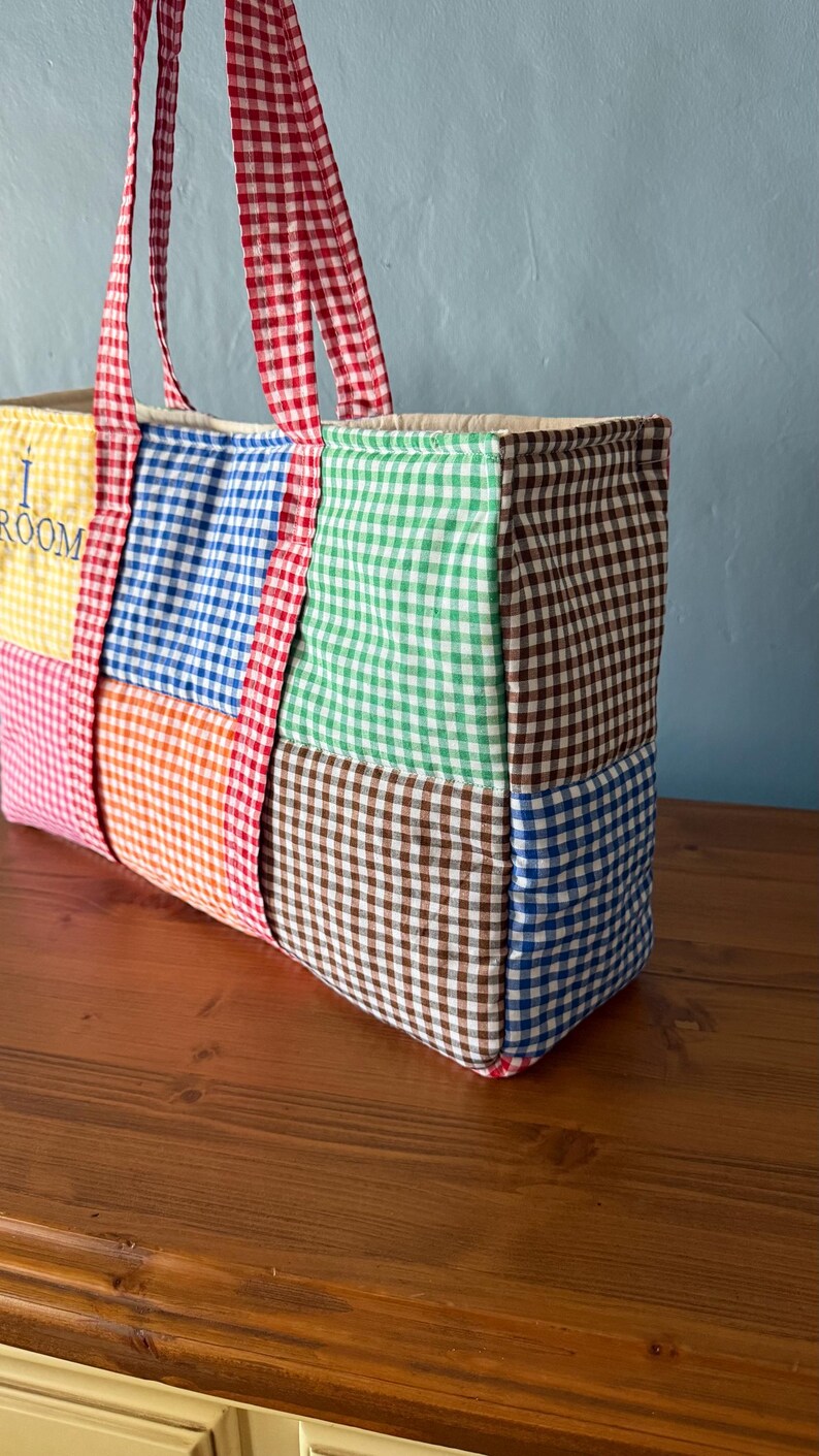 Puede incluir: Un bolso tote rectangular hecho de paneles de tela acolchada con un patr&oacute;n de cuadros vichy multicolor en amarillo, azul, naranja, verde y marr&oacute;n. El bolso tiene dos asas de cuadros vichy rojos y blancos y el texto I ROOM.