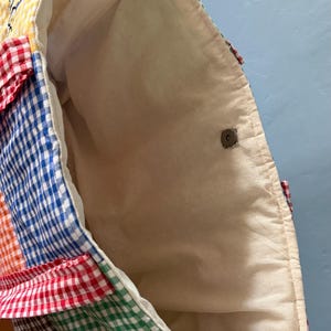 Puede incluir: Un bolso tote con un dise&ntilde;o de patchwork de cuadrados de tela de cuadros vichy multicolor en amarillo, rojo, azul, naranja, verde y marr&oacute;n. El interior est&aacute; forrado con tela color hueso e incluye un cierre magn&eacute;tico de tono plateado.