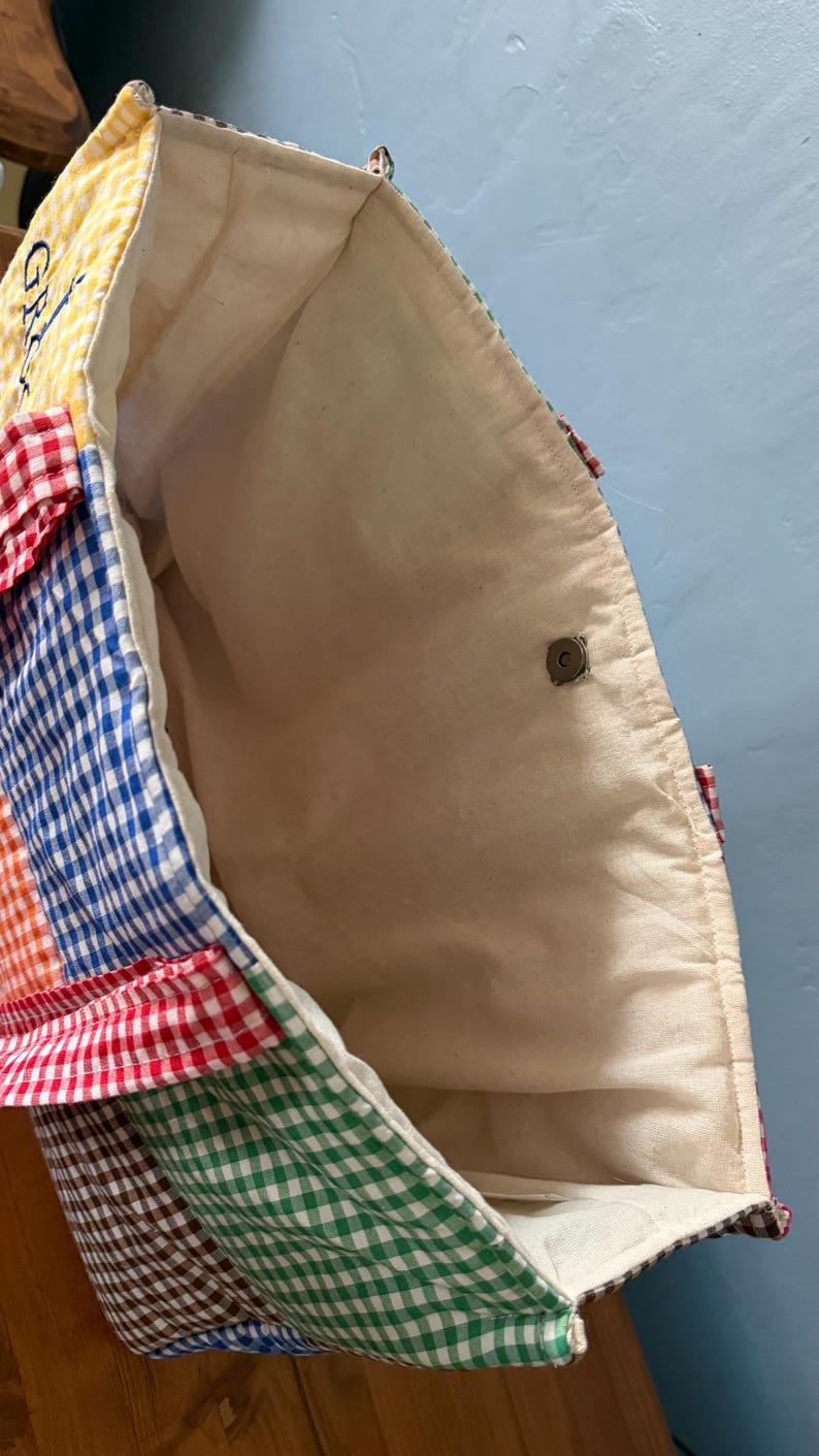 Puede incluir: Un bolso tote con un dise&ntilde;o de patchwork de cuadrados de tela de cuadros vichy multicolor en amarillo, rojo, azul, naranja, verde y marr&oacute;n. El interior est&aacute; forrado con tela color hueso e incluye un cierre magn&eacute;tico de tono plateado.