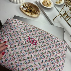 Puede incluir: Una funda para portátil con estampado floral de flores rosas y azules sobre fondo blanco. Las iniciales "NB" están bordadas en rosa. Pendientes dorados expuestos en platos decorativos.