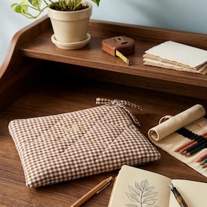 Può includere: Una pochette rettangolare trapuntata con motivo a quadretti vichy marrone e bianco, caratterizzata dalle iniziali "MI" ricamate sul davanti. La pochette presenta una chiusura a laccio in tessuto sull'angolo superiore ed è realizzata in tessuto imbottito.