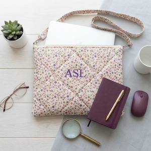 Könnte beinhalten: Eine gesteppte Laptoptasche aus cremefarbenem Stoff mit einem kleinen Blumenmuster in Lila-, Rosa- und Grüntönen. Die Tasche verfügt über einen passenden Schulterriemen und ist in der Mitte mit den violetten Monogrammbuchstaben ASL bestickt.