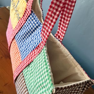 Puede incluir: Un bolso tote de patchwork confeccionado con cuadrados de tela de cuadros vichy multicolor en amarillo, rosa, naranja, azul, verde y marr&oacute;n. El bolso cuenta con asas de cuadros rojos y blancos y la palabra GROOM bordada en azul en el panel amarillo.