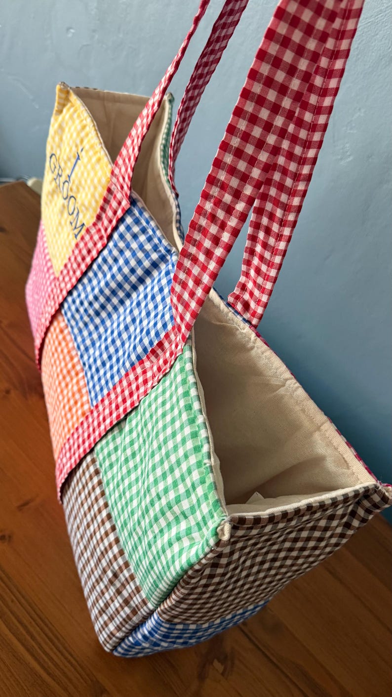 Puede incluir: Un bolso tote de patchwork confeccionado con cuadrados de tela de cuadros vichy multicolor en amarillo, rosa, naranja, azul, verde y marr&oacute;n. El bolso cuenta con asas de cuadros rojos y blancos y la palabra GROOM bordada en azul en el panel amarillo.