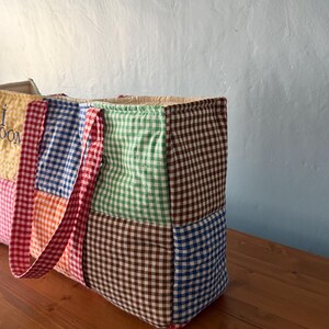 Puede incluir: Un bolso tote acolchado rectangular construido con varios cuadrados de tela de cuadros vichy en colores que incluyen marr&oacute;n, verde, azul, naranja y amarillo. El bolso cuenta con dos asas de cuadros rojos y blancos y un panel amarillo parcial con texto.