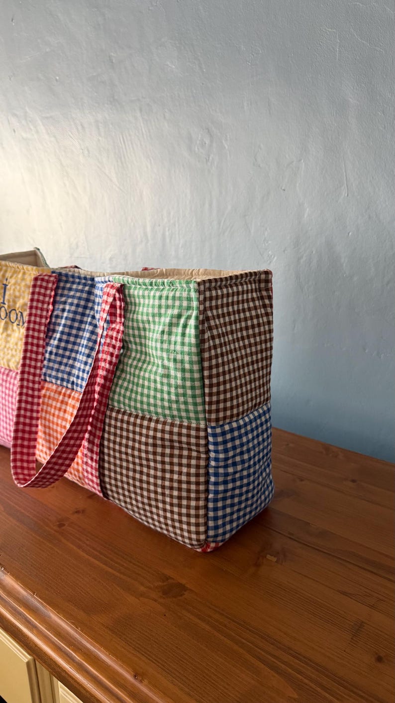Puede incluir: Un bolso tote acolchado rectangular construido con varios cuadrados de tela de cuadros vichy en colores que incluyen marr&oacute;n, verde, azul, naranja y amarillo. El bolso cuenta con dos asas de cuadros rojos y blancos y un panel amarillo parcial con texto.
