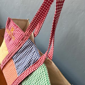 Puede incluir: Un bolso tote de patchwork con un patr&oacute;n de cuadros vichy multicolor en cuadrados rojos, amarillos, naranjas, azules, verdes y marrones. El bolso tiene dos asas de cuadros rojos y blancos e incluye el texto bordado en amarillo BRIDE.