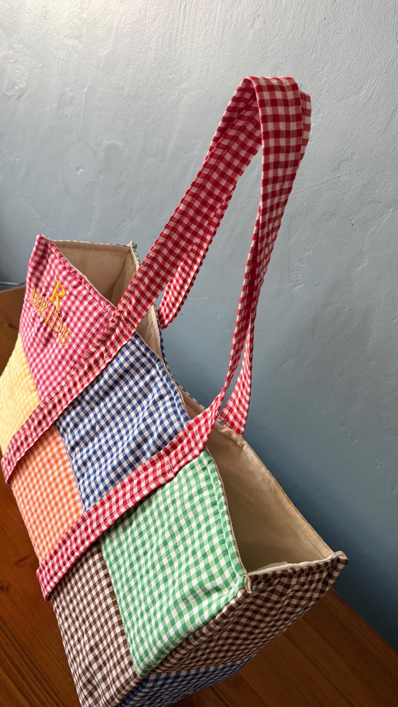 Puede incluir: Un bolso tote de patchwork con un patr&oacute;n de cuadros vichy multicolor en cuadrados rojos, amarillos, naranjas, azules, verdes y marrones. El bolso tiene dos asas de cuadros rojos y blancos e incluye el texto bordado en amarillo BRIDE.
