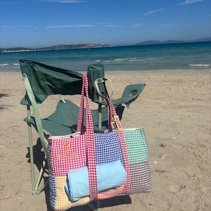 Puede incluir: Un bolso rectangular con dise&ntilde;o de patchwork de cuadros vichy en rojo, azul, verde, amarillo y marr&oacute;n. El bolso tiene asas de cuadros rojos y una letra R bordada en la parte delantera. Un objeto de tela azul claro est&aacute; metido en el frente.
