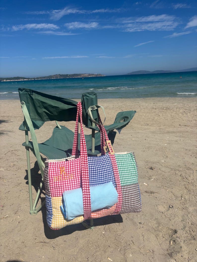Puede incluir: Un bolso rectangular con dise&ntilde;o de patchwork de cuadros vichy en rojo, azul, verde, amarillo y marr&oacute;n. El bolso tiene asas de cuadros rojos y una letra R bordada en la parte delantera. Un objeto de tela azul claro est&aacute; metido en el frente.