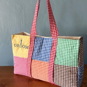 Puede incluir: Un bolso tote rectangular con un dise&ntilde;o de patchwork de cuadros vichy multicolores en amarillo, azul, verde, naranja, rosa y marr&oacute;n. El bolso tiene dos asas de cuadros vichy rojos y blancos y el texto I GROOM bordado en azul oscuro.