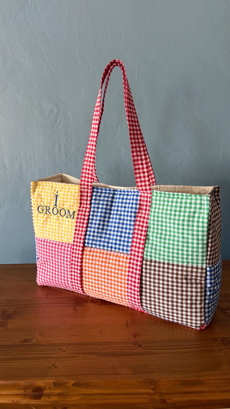 Puede incluir: Un bolso tote rectangular con un dise&ntilde;o de patchwork de cuadros vichy multicolores en amarillo, azul, verde, naranja, rosa y marr&oacute;n. El bolso tiene dos asas de cuadros vichy rojos y blancos y el texto I GROOM bordado en azul oscuro.
