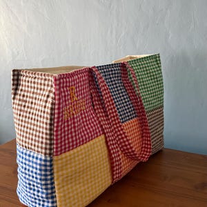 Puede incluir: Un bolso tote rectangular confeccionado con un patchwork de cuadrados de tela de cuadros vichy multicolor en marr&oacute;n, rojo, azul, amarillo, naranja y verde. La secci&oacute;n roja presenta el texto amarillo bordado R BRIDE. Tiene dos asas de vichy a juego.