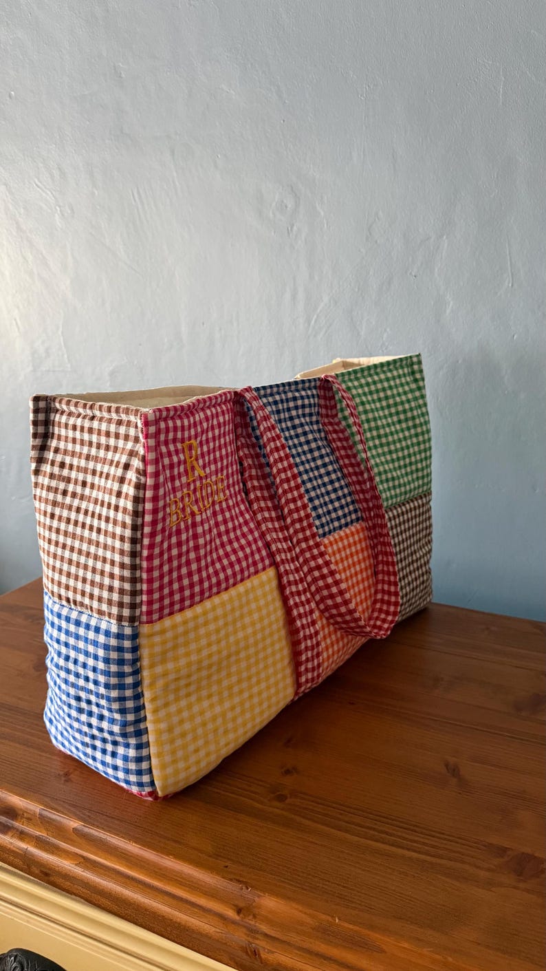 Puede incluir: Un bolso tote rectangular confeccionado con un patchwork de cuadrados de tela de cuadros vichy multicolor en marr&oacute;n, rojo, azul, amarillo, naranja y verde. La secci&oacute;n roja presenta el texto amarillo bordado R BRIDE. Tiene dos asas de vichy a juego.