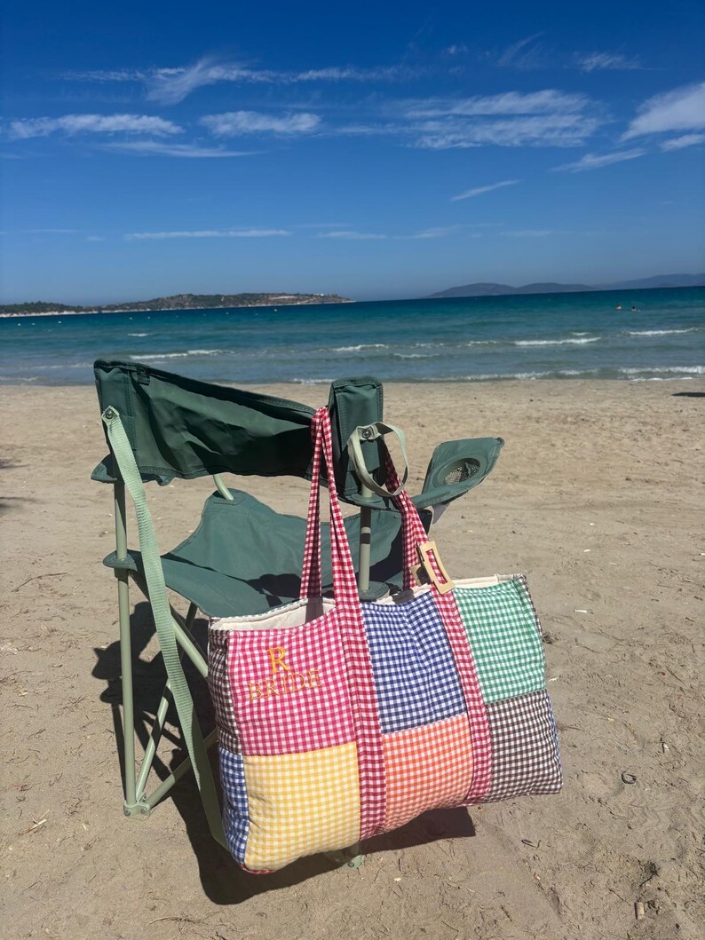 Puede incluir: Un bolso tote rectangular grande con un dise&ntilde;o de patchwork de cuadros vichy multicolores en rojo, azul, verde, amarillo y naranja. El bolso tiene dos asas largas de tela y la letra R bordada con hilo dorado en la secci&oacute;n roja.