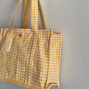 Może przedstawiać: Żółto-biała torba typu tote w kratę z pasującymi paskami i małym pomarańczowym zapięciem na guzik. Torba ma prostokątny kształt i wisi na białej ścianie. Etykieta brzmi "bonne maman".