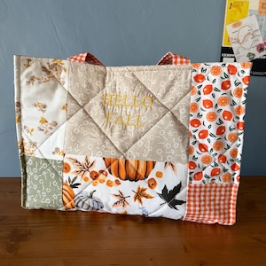 May include: A quilted tote bag featuring a patchwork design with beige floral fabric, a white panel with orange pumpkins and autumn leaves, and a side panel with orange citrus fruit prints. The text HELLO FALL is embroidered in gold thread.