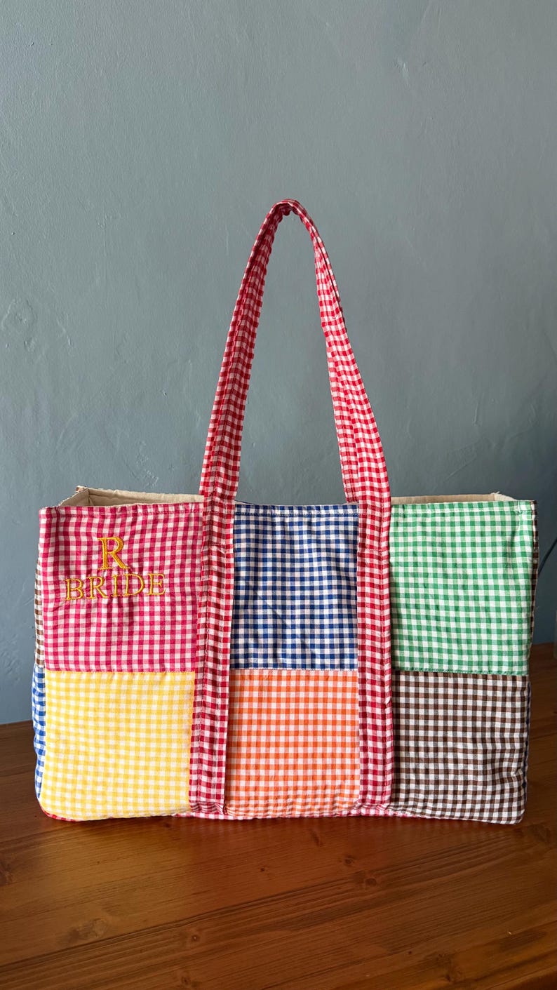 Puede incluir: Un bolso tote rectangular con un patr&oacute;n de patchwork de cuadros vichy multicolor en rojo, azul, verde, amarillo, naranja y marr&oacute;n. El bolso tiene dos asas de cuadros vichy rojos y blancos y el texto bordado en amarillo R BRIDE en el frente.