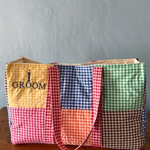 Puede incluir: Un bolso tote rectangular confeccionado con paneles de tela acolchada que presentan un patr&oacute;n de cuadros vichy multicolor en amarillo, azul, naranja, verde, marr&oacute;n y rosa. El panel amarillo muestra el texto negro bordado I GROOM.