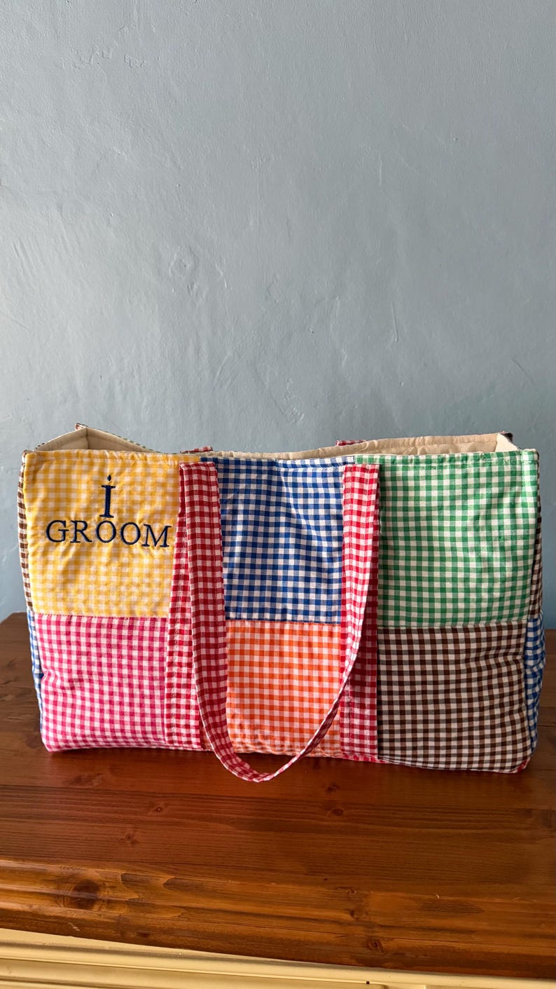 Puede incluir: Un bolso tote rectangular confeccionado con paneles de tela acolchada que presentan un patr&oacute;n de cuadros vichy multicolor en amarillo, azul, naranja, verde, marr&oacute;n y rosa. El panel amarillo muestra el texto negro bordado I GROOM.