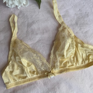 May include: A pale yellow bra with delicate lace detailing on the cups and straps. The bra features a small bow at the center and adjustable straps. The soft, satin-like material gives it a vintage aesthetic.