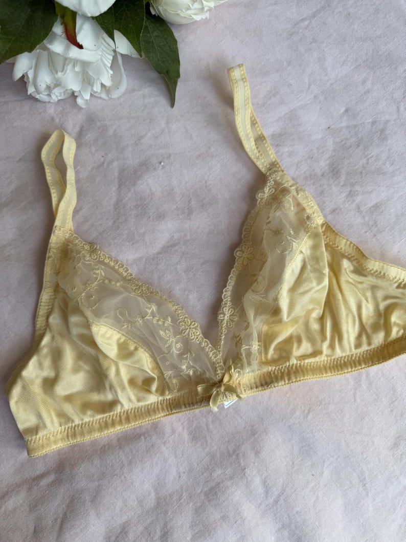 May include: A pale yellow bra with delicate lace detailing on the cups and straps. The bra features a small bow at the center and adjustable straps. The soft, satin-like material gives it a vintage aesthetic.
