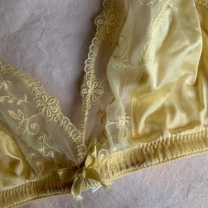 May include: A pale yellow bra with delicate floral lace detailing. The bra features a soft, silky fabric and a small bow at the center. The design includes intricate embroidery and a scalloped edge.