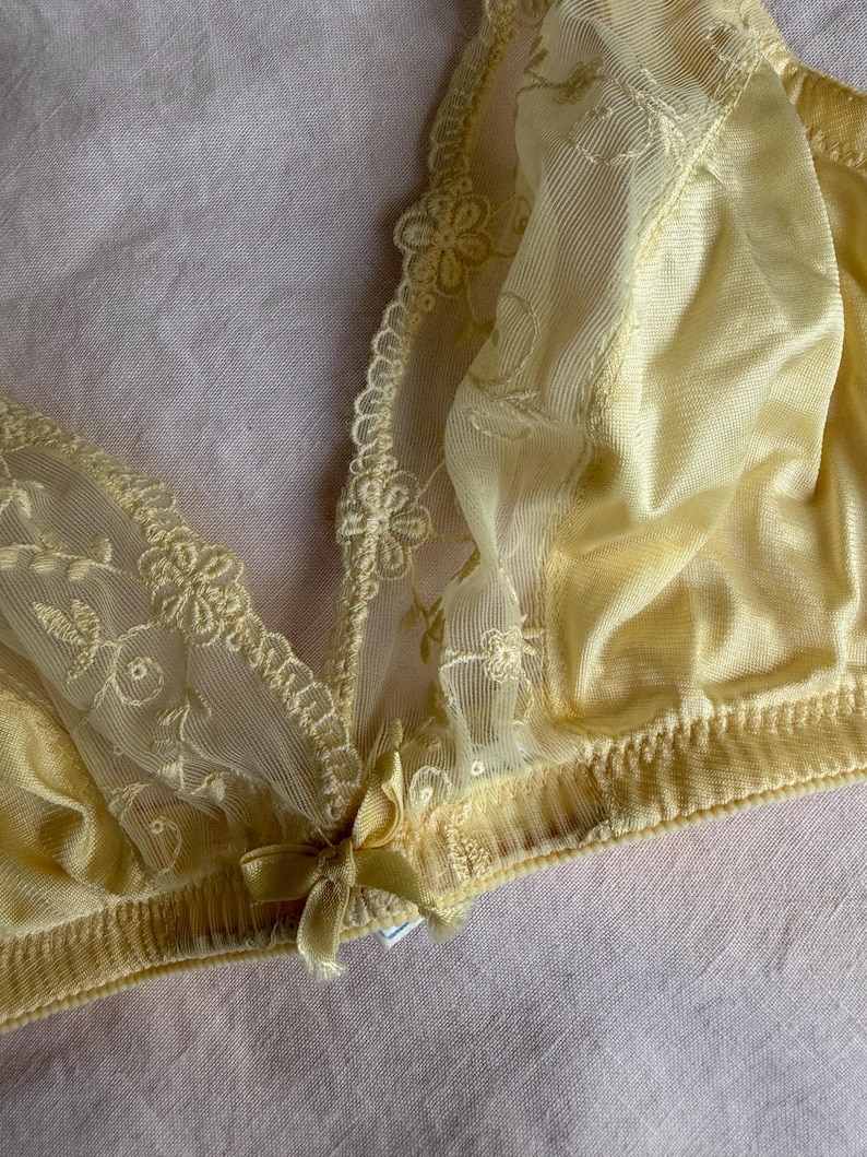 May include: A pale yellow bra with delicate floral lace detailing. The bra features a soft, silky fabric and a small bow at the center. The design includes intricate embroidery and a scalloped edge.