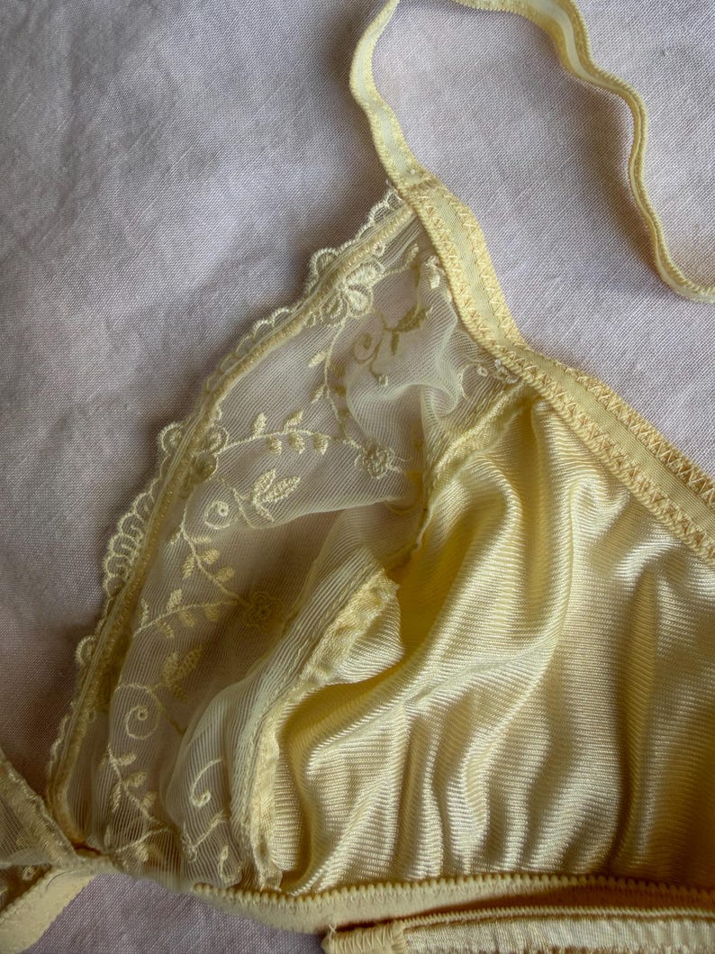 May include: A pale yellow bra with delicate lace detailing. The bra features a soft, silky fabric with a subtle sheen. The straps and edges are trimmed with a matching yellow color, creating a cohesive and elegant design.