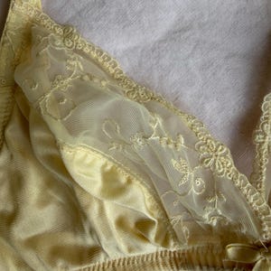 May include: A pale yellow bra with delicate lace detailing. The bra features a soft, silky fabric and intricate floral embroidery on the sheer lace panels. The edges are trimmed with scalloped lace, and a small bow adorns the front.