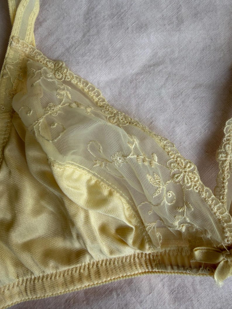May include: A pale yellow bra with delicate lace detailing. The bra features a soft, silky fabric and intricate floral embroidery on the sheer lace panels. The edges are trimmed with scalloped lace, and a small bow adorns the front.