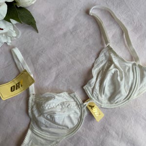 May include: A white bra with lace trim and adjustable straps. The bra has a tag that reads "ORY". The bra is made of a soft, smooth fabric and is displayed on a pink surface.