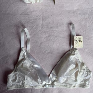 May include: A white triangle bra with adjustable straps. The bra has a satin-like finish and a small bow detail at the center. A tag that says "Little K" is attached to the strap. The bra is set against a pink background with a white flower.