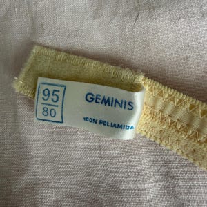 May include: Close-up of a pale yellow bra strap with a label. The label reads "GEMINIS" and "100% POLIAMIDA" with size markings "95" and "80". The strap is on a pink fabric background.